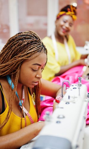 Two african dressmaker woman sews clothes on sewing machine at tailor office. Black seamstress girls.